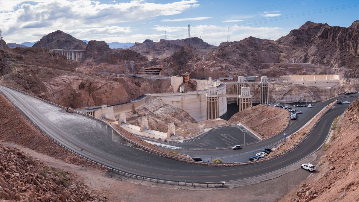 A wide panorama of a curving paved road winding down through reddish rocky canyon walls toward a large concrete structure with towers.