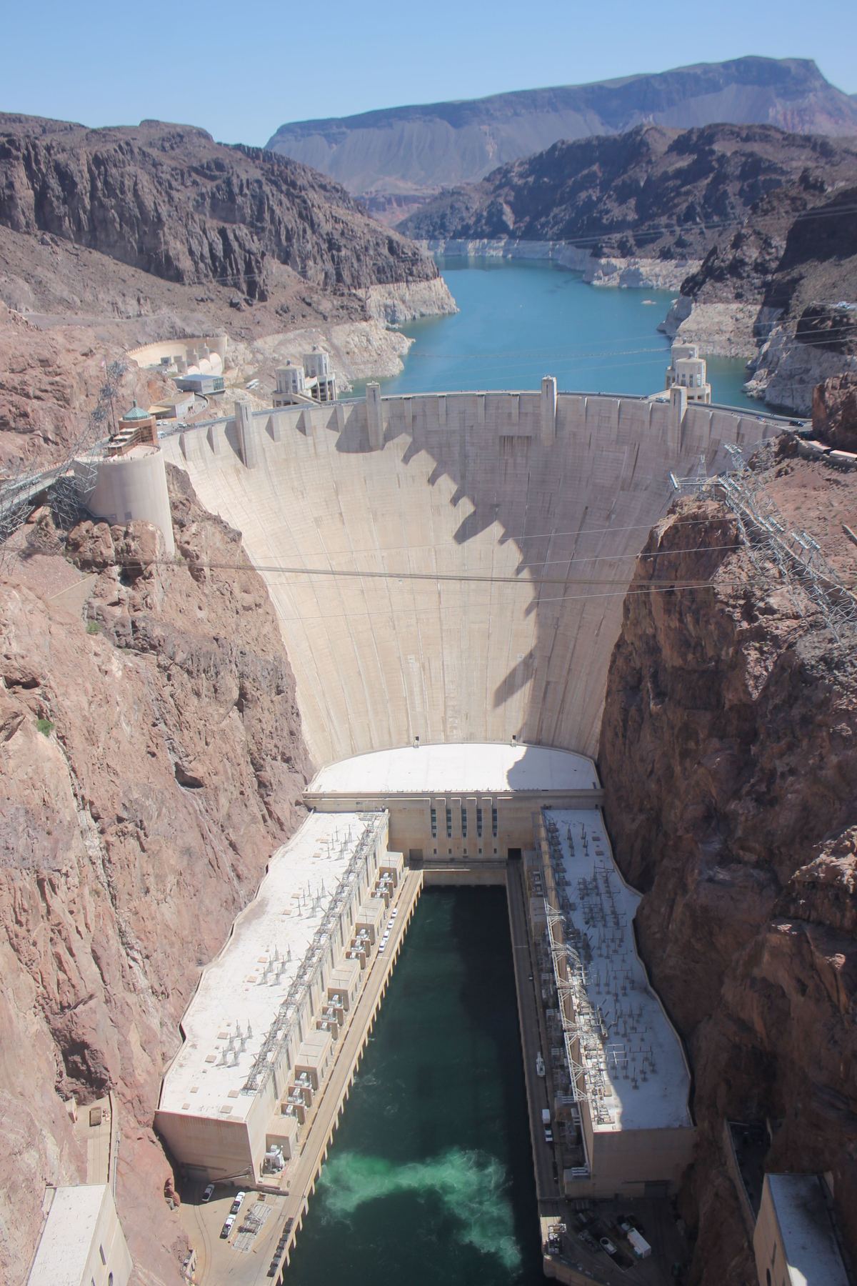 A curved concrete dam between rust-colored canyon walls, with a blue lake behind it and a power plant beside green water below.