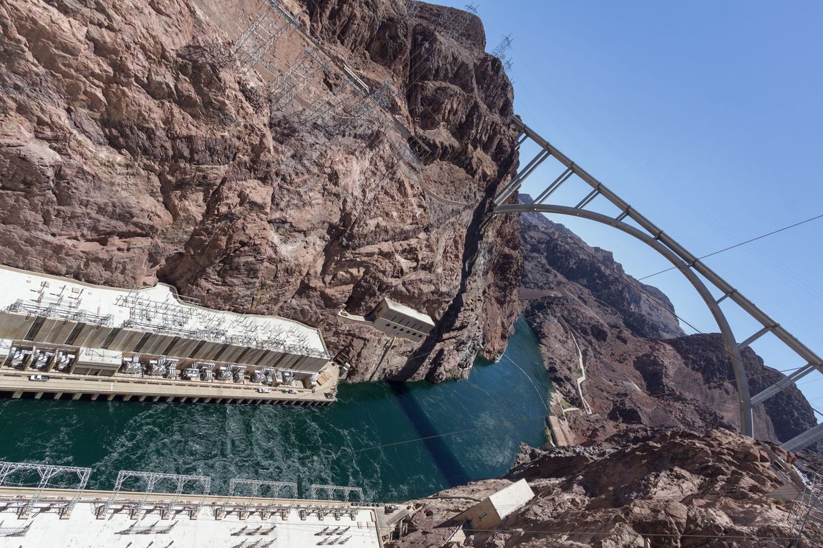 Looking down a rugged red-brown canyon at power plant buildings beside green river water, with a steel arch bridge curving across the blue sky.