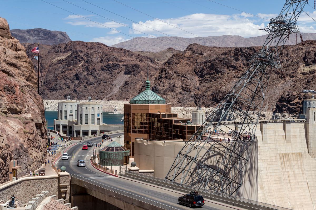 A road curves past a dam wall, a steel power tower and buildings with green roofs, with rugged brown hills and blue water behind.