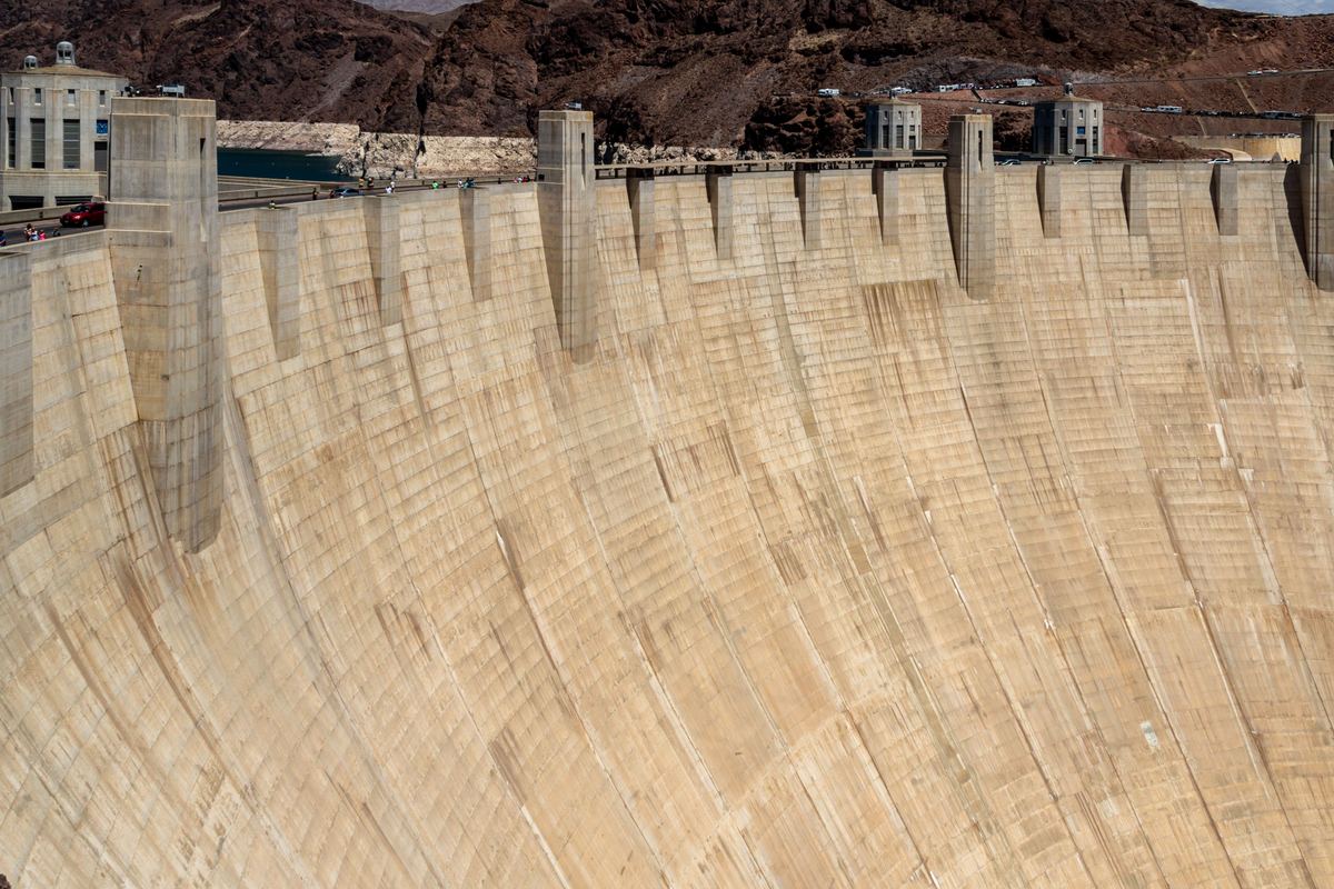 A huge curved concrete dam wall with tall intake towers along the top, set against dry reddish-brown rocky hills.