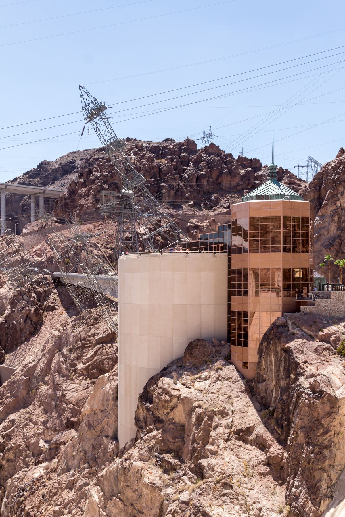 A tall cylindrical concrete tower and a copper-glass tower sit against a rocky canyon wall, with electricity pylons and power lines under a clear blue sky.